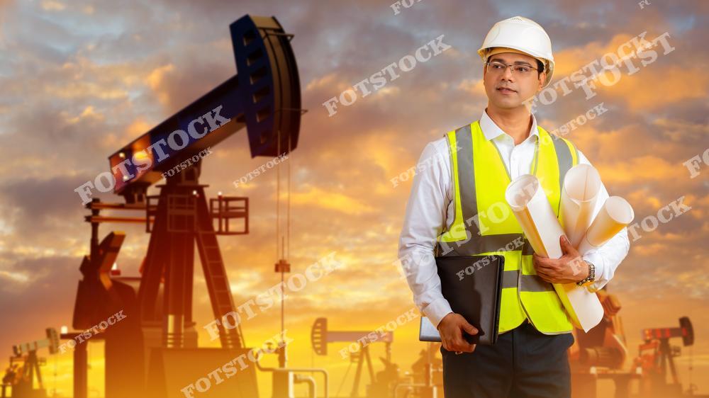Мужчина в защитном шлеме и жилете держит чертежи на фоне нефтяных вышек. Он одет в рабочую униформу и стоит при закате, что создаёт драматичное и вдохновляющее ощущение. Его выражение лица серьезное и внимательное, он кажется профессионалом в своей области. Небо за его спиной оранжево-голубое, добавляя контраст и глубину композиции.
